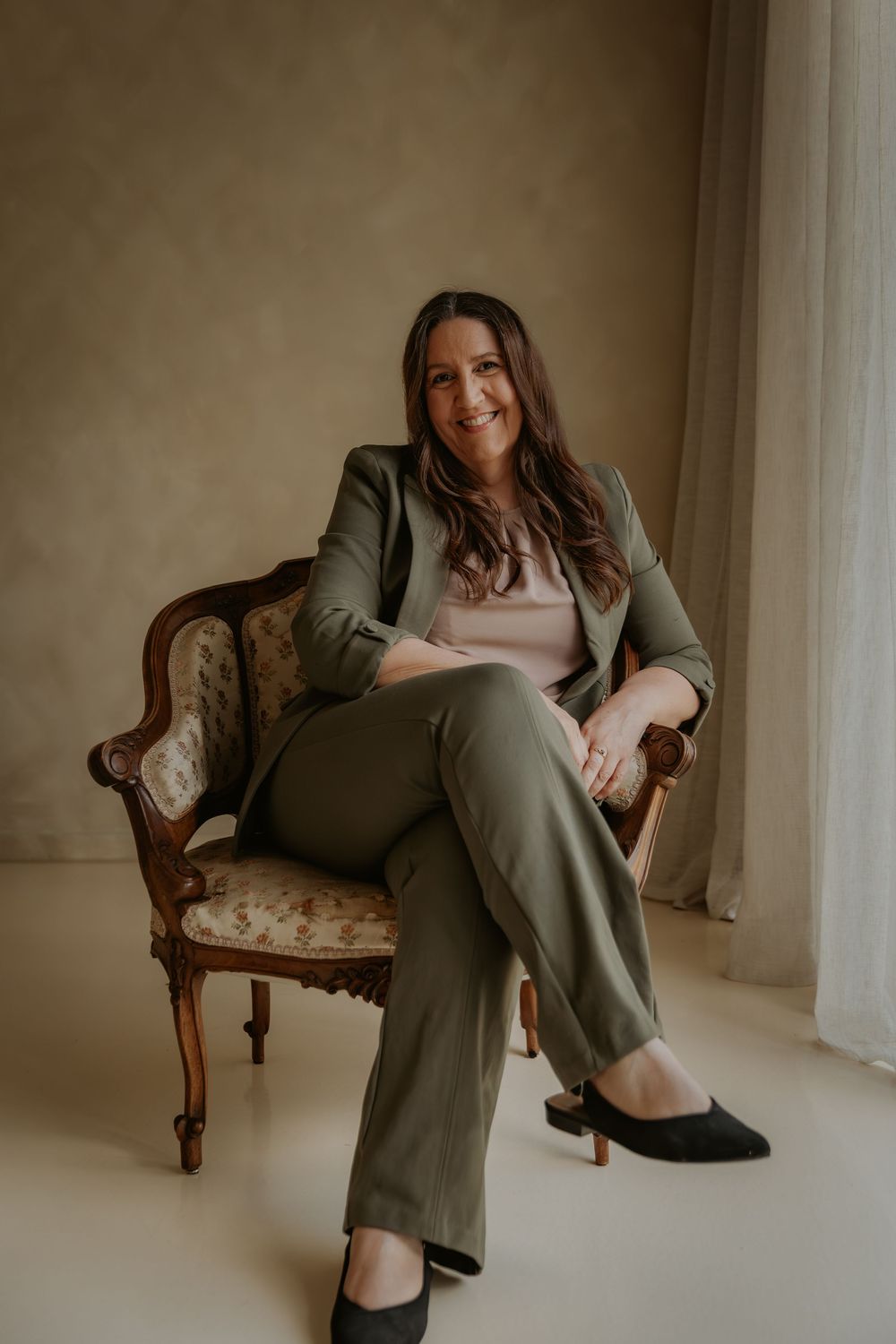 Woman in olive green suit sitting confidently in ornate wooden chair, smiling warmly in neutral toned studio.