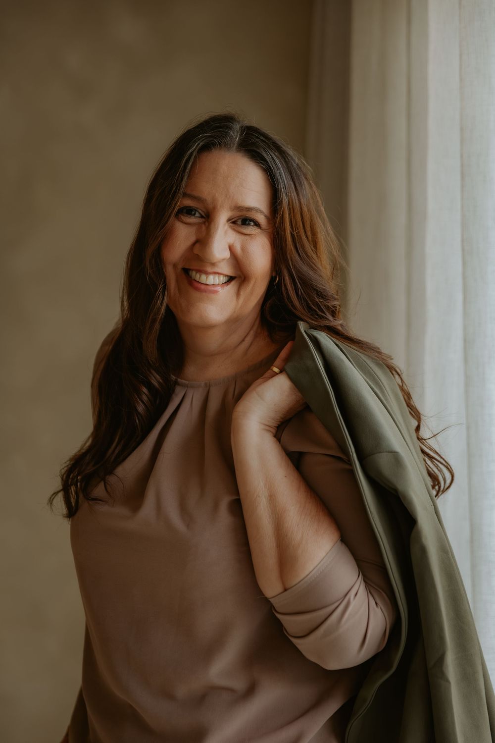 Smiling woman with brown hair holding a green jacket over her shoulder near a bright window.