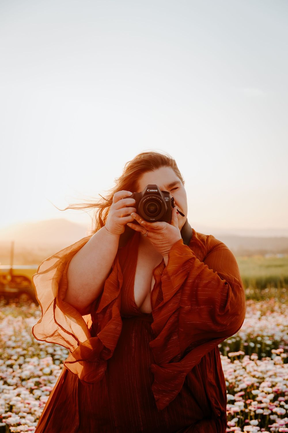 A photographer in a flowing rust-colored dress takes pictures in a flower field at golden hour sunset.