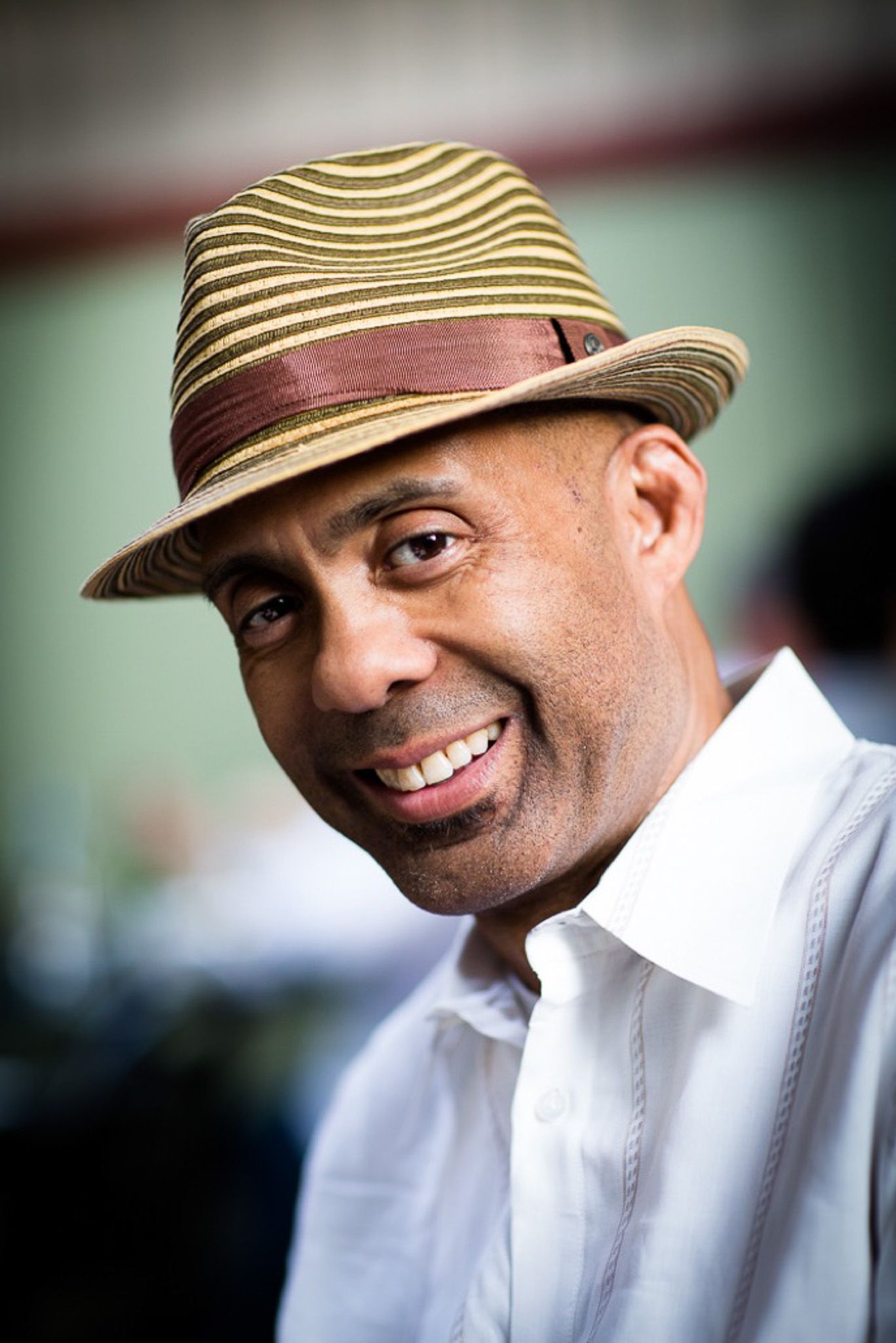 A smiling man wearing a striped straw fedora hat and white shirt outdoors.