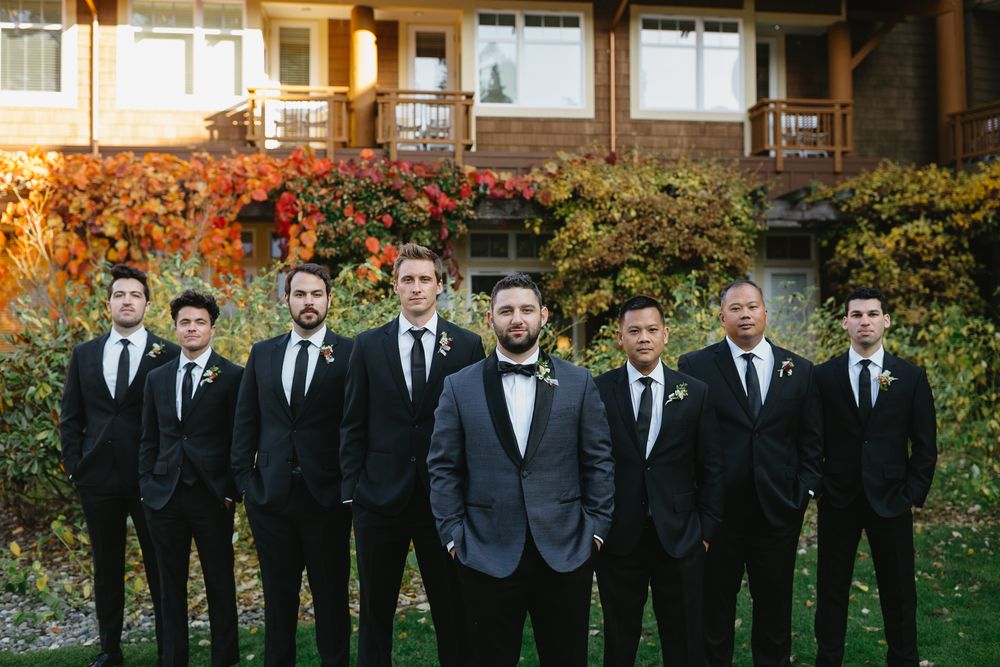 A wedding party in black suits and ties stands in a row on grass with autumn foliage in the background.