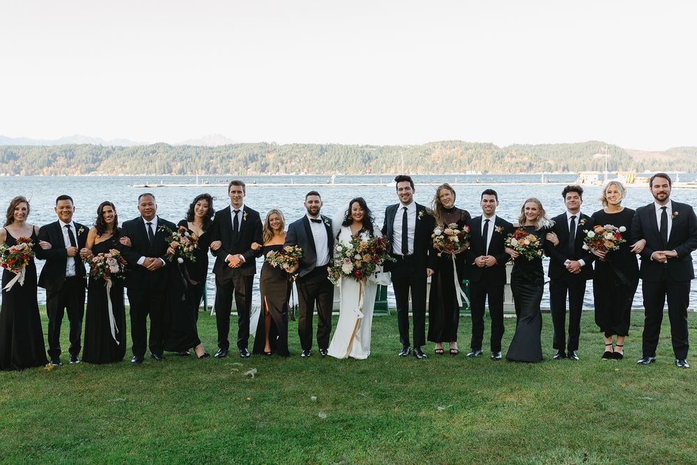 Wedding party wearing black attire stands in a line on green grass against a scenic coastal backdrop.
