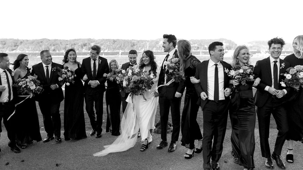 A wedding party in black and white walks together in a line outdoors, with the bride in a flowing white dress.