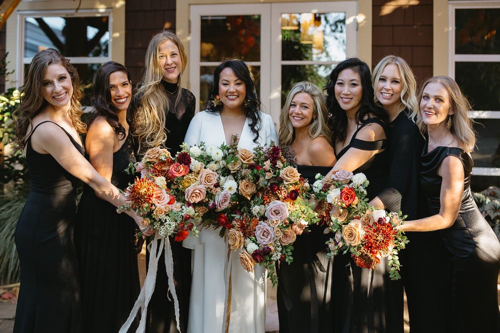 Bridesmaids in black dresses holding vibrant orange and pink floral bouquets pose together at an outdoor wedding.