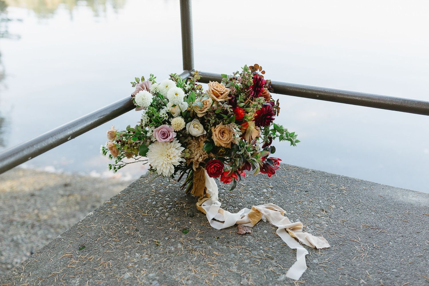 Wedding bouquet with pink and orange flowers tied with white ribbon rests on concrete ledge outdoors.