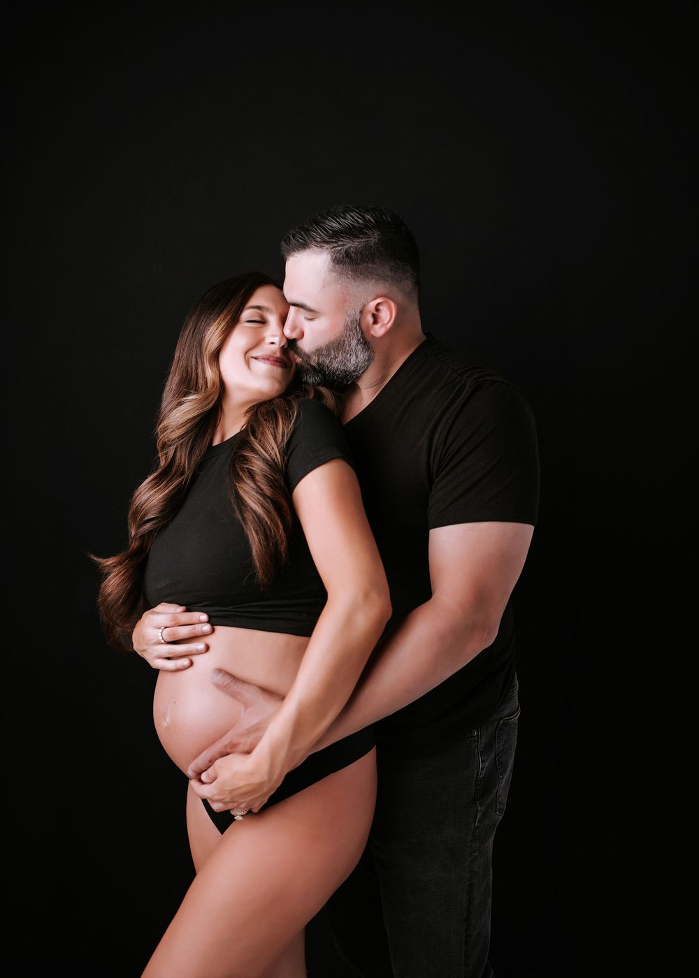 Couple in black outfits sharing a tender maternity photo moment against a dark studio background.