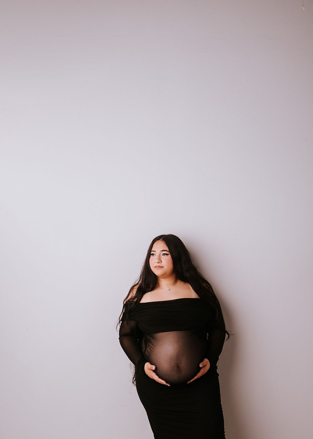 Pregnant woman in black off-shoulder maternity gown cradling baby bump against white studio background.