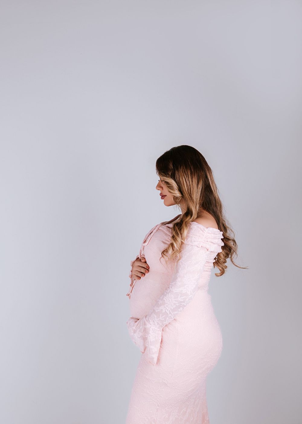 Maternity portrait of woman in pink lace dress against grey background.