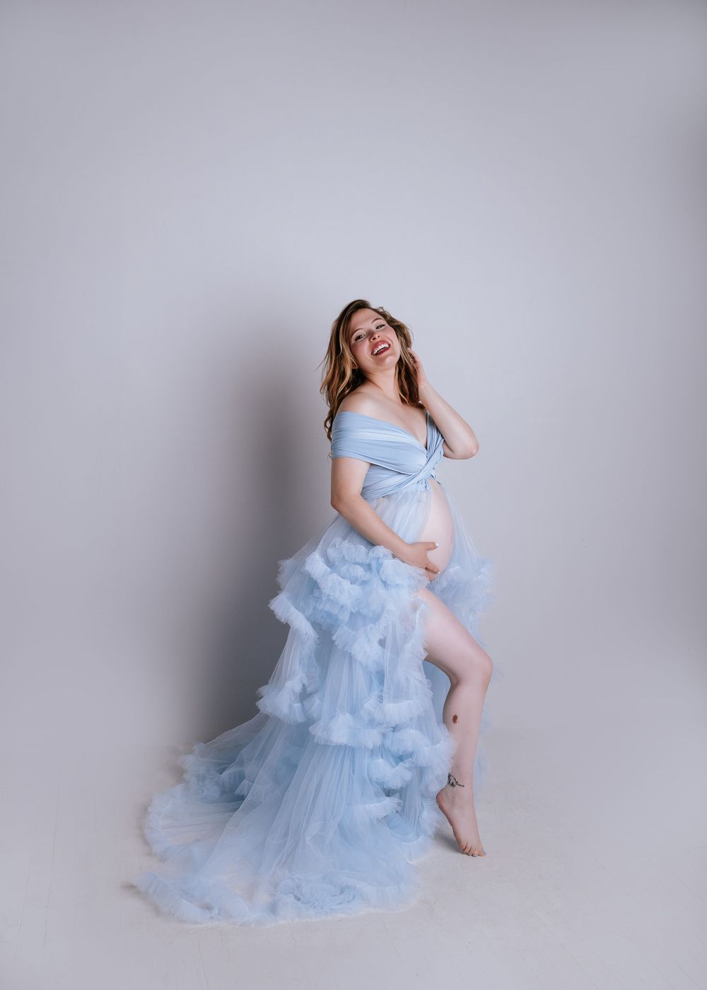 Pregnant woman in a light blue ruffled tulle maternity gown smiling joyfully in a studio setting.
