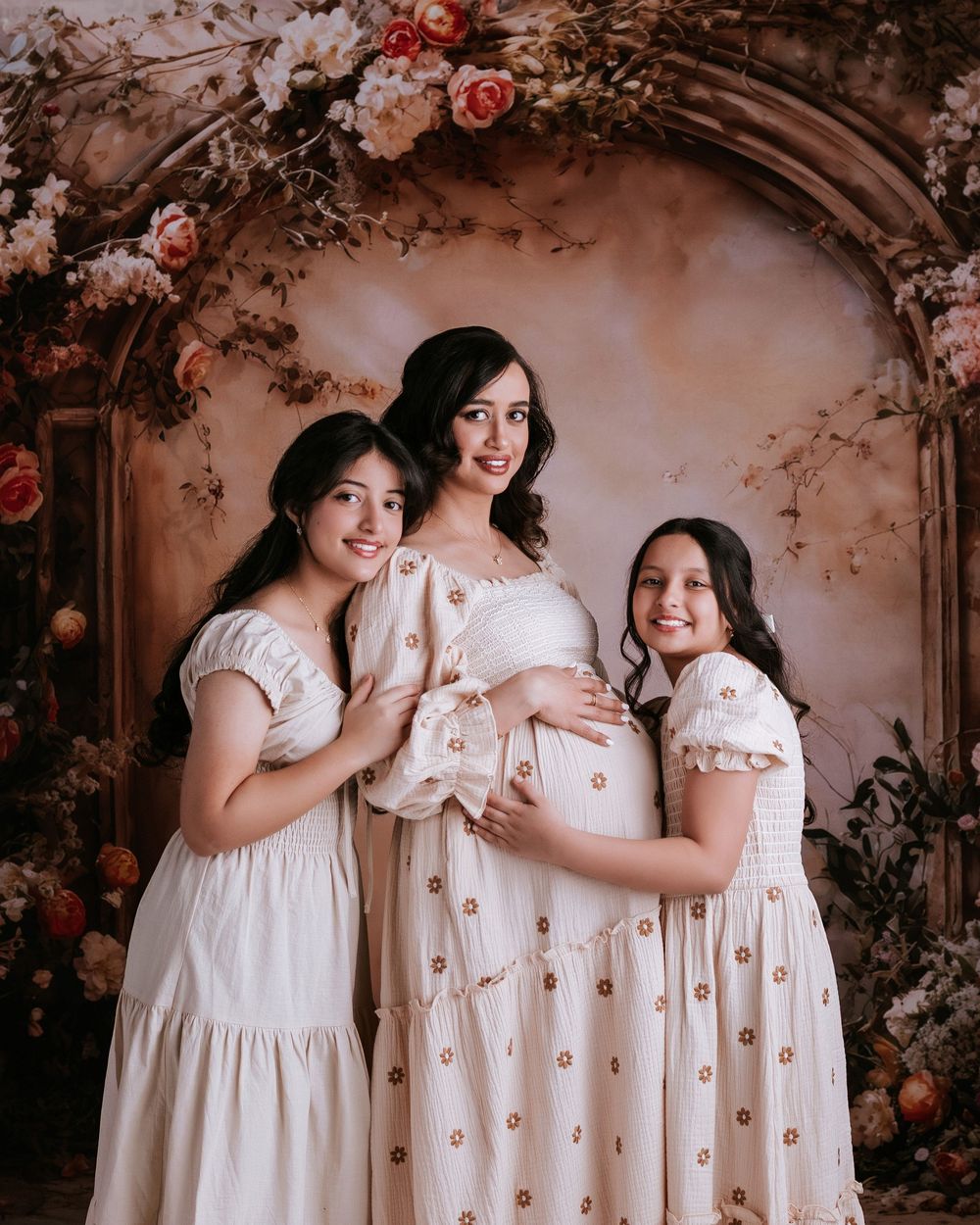 Pregnant woman in white dress poses with two girls in floral studio backdrop for maternity photo.