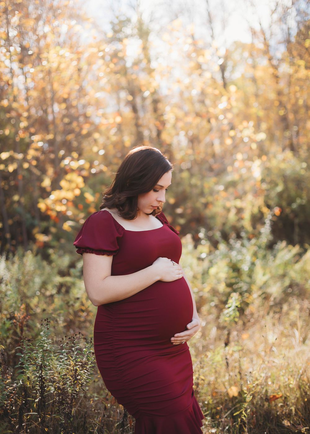 Expectant mother in burgundy dress standing in autumn forest setting.