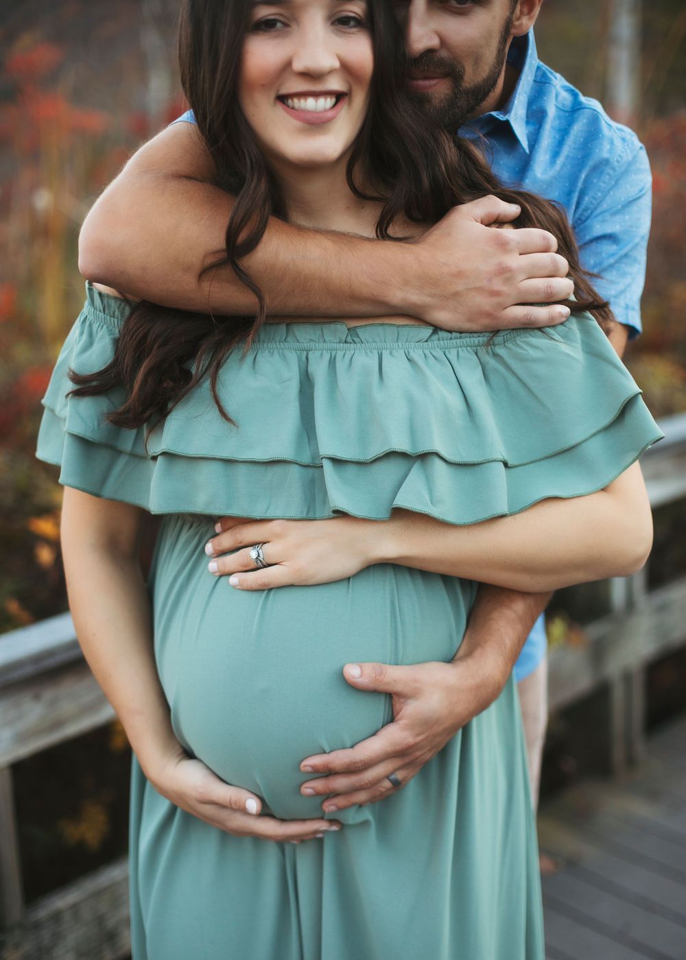 A couple embraces in front of autumn foliage wearing a mint green off-shoulder dress and dark casual attire.