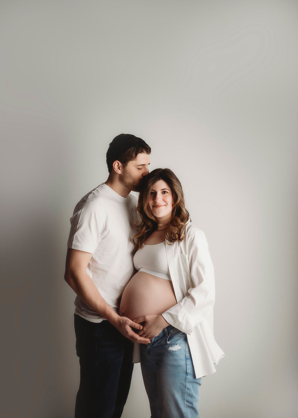 Intimate maternity studio session with couple in white and denim against grey background.