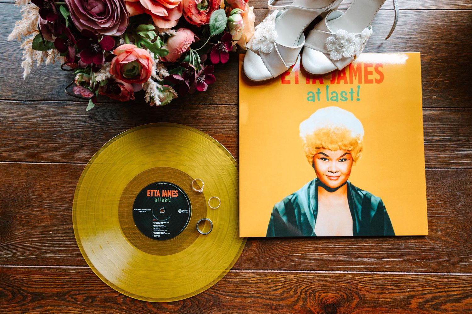 A yellow vinyl record and orange album cover displayed on wooden surface with roses and sandals in the background.