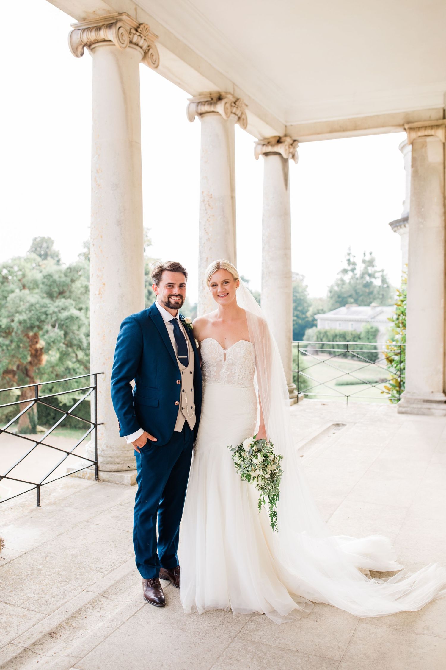 Elegant wedding couple shares romantic moments under white columned porch in natural sunlight.