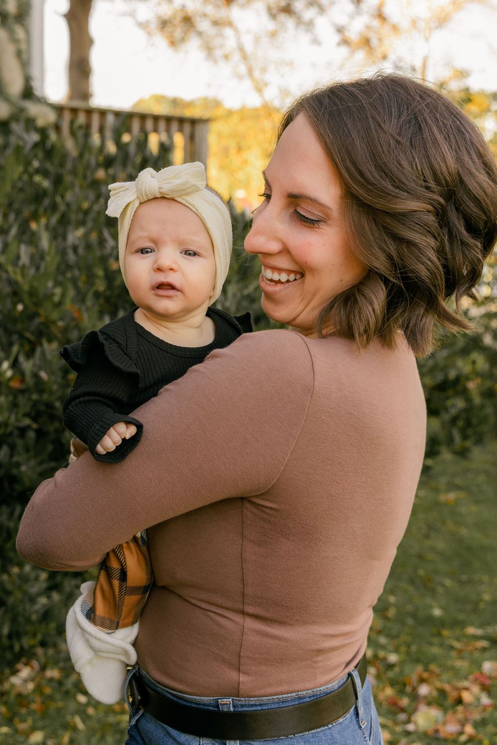 Parent wearing brown sweater holds baby in white outfit during autumn outdoor photoshoot with fallen leaves.