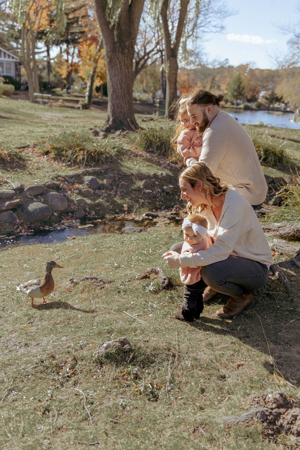 A family feeds a duck by the water on an autumn day in the park with fall foliage and trees in the background.