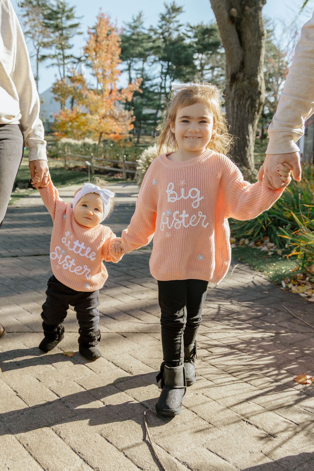 Four siblings wearing matching pink sweaters with text stand in a line on a stone path during autumn.