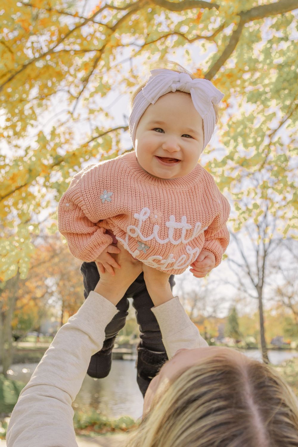 A joyful moment captured in autumn as a small child in a pink sweater and hat is lifted up against golden fall foliage.