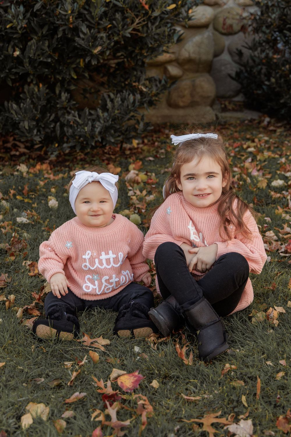Two children in matching pink sweaters sit together on grass surrounded by autumn leaves.