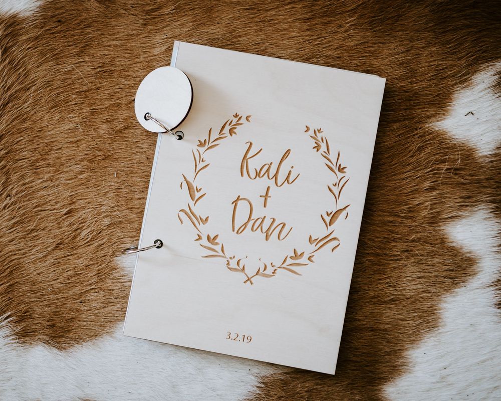Wooden wedding guest book engraved with bride and groom names Kali and Dan with a laurel wreath design.