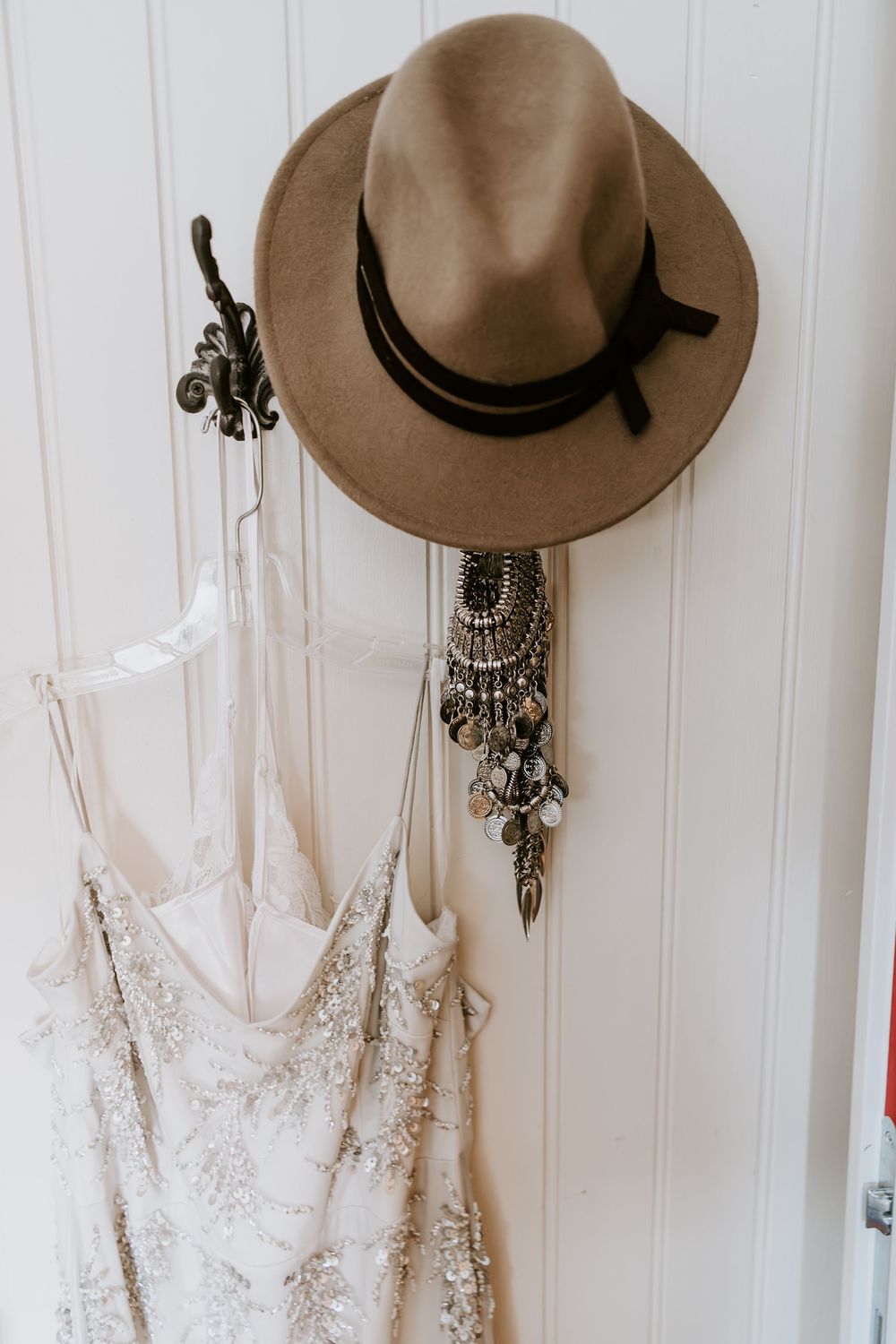 Boho bridal accessories including a felt fedora, beaded gown, and statement jewelry hanging on a white door.