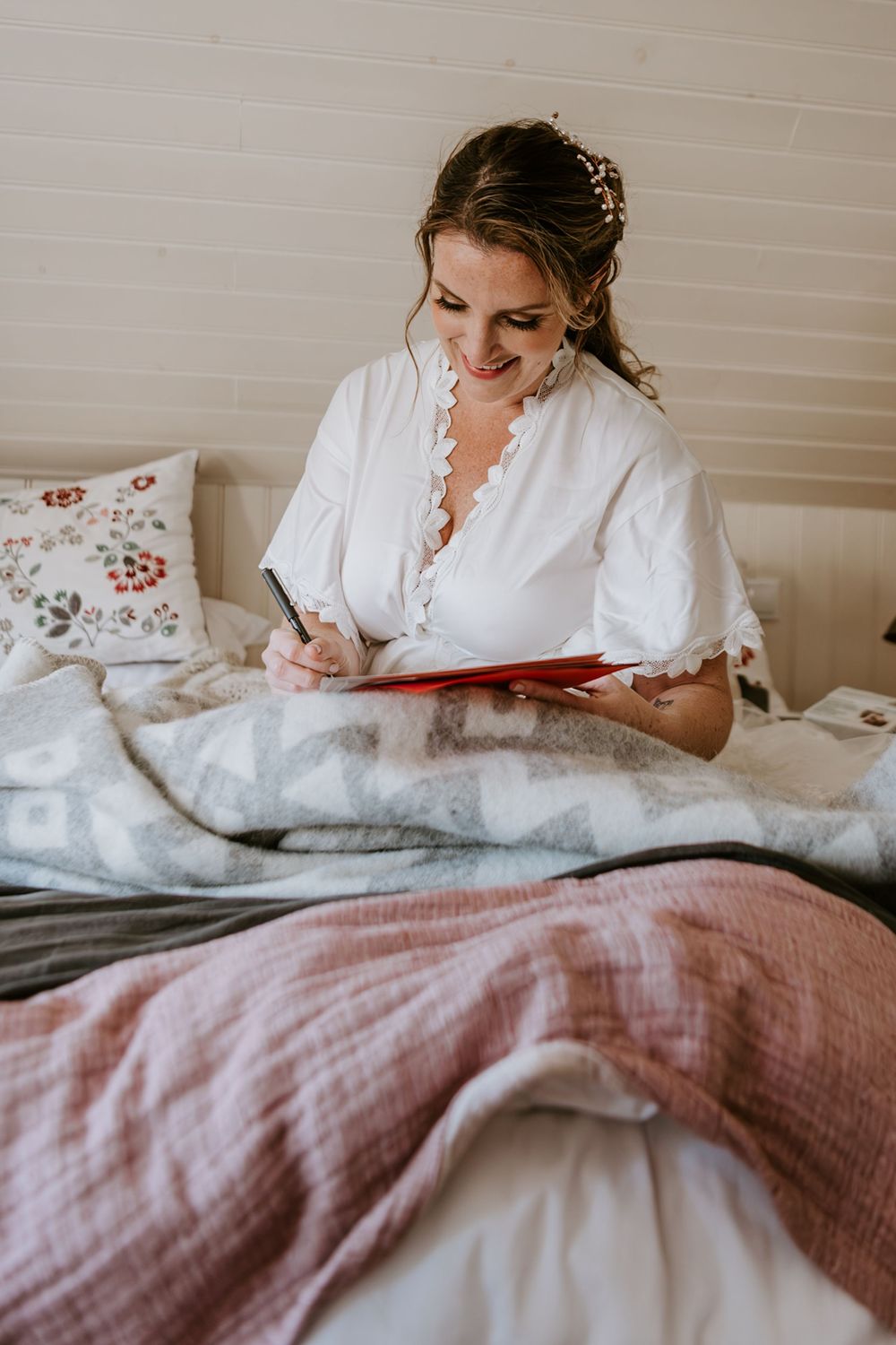 Smiling bride in white robe writing vows in a red notebook while sitting in a cozy bed.
