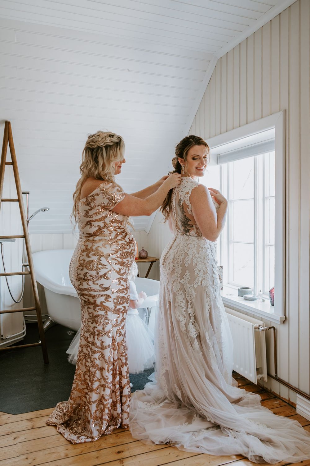 Bridesmaid in floral gown helping bride fasten her lace wedding dress near a clawfoot bathtub.