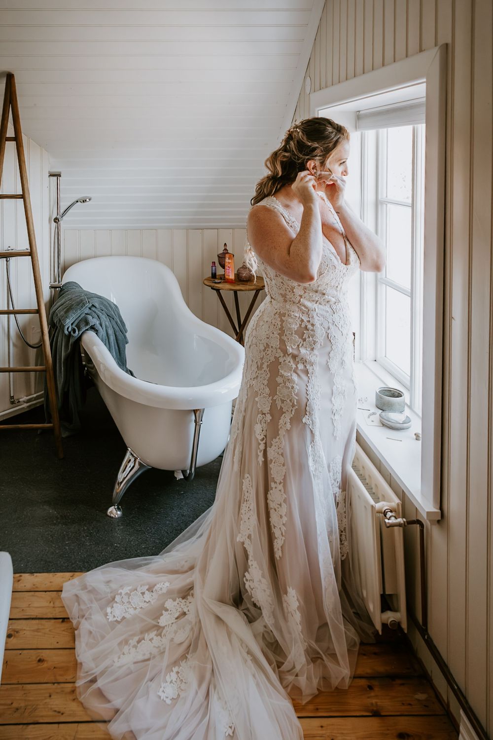 Bride in stunning lace wedding gown with long train putting on earrings by a sunlit window.