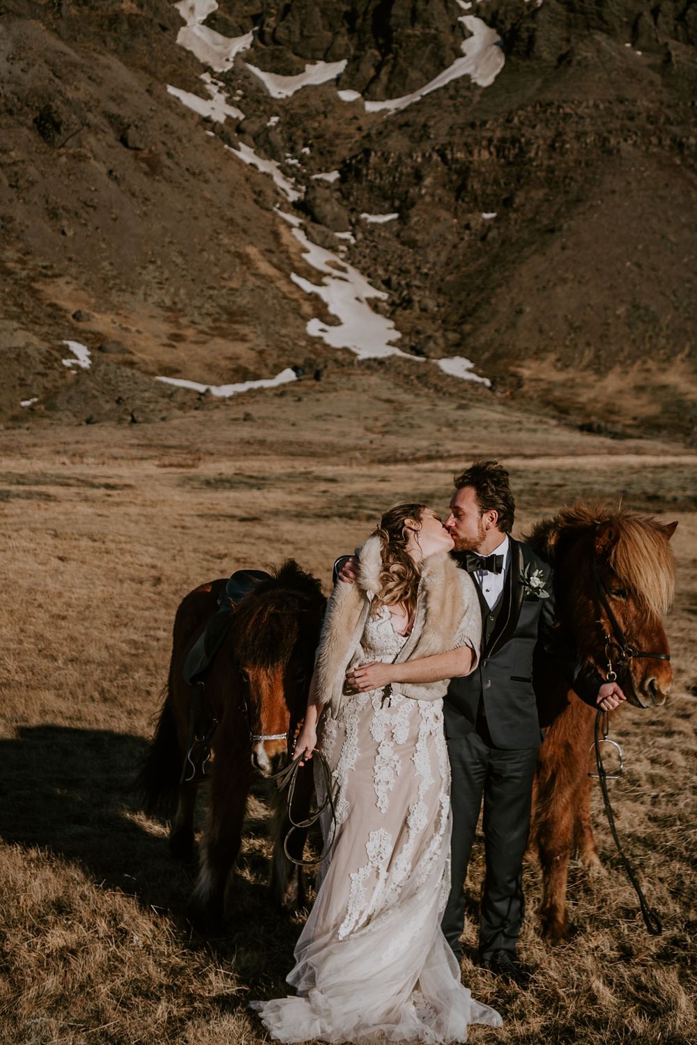 Bride and groom share a kiss surrounded by Icelandic horses with snow-capped mountains in the background.