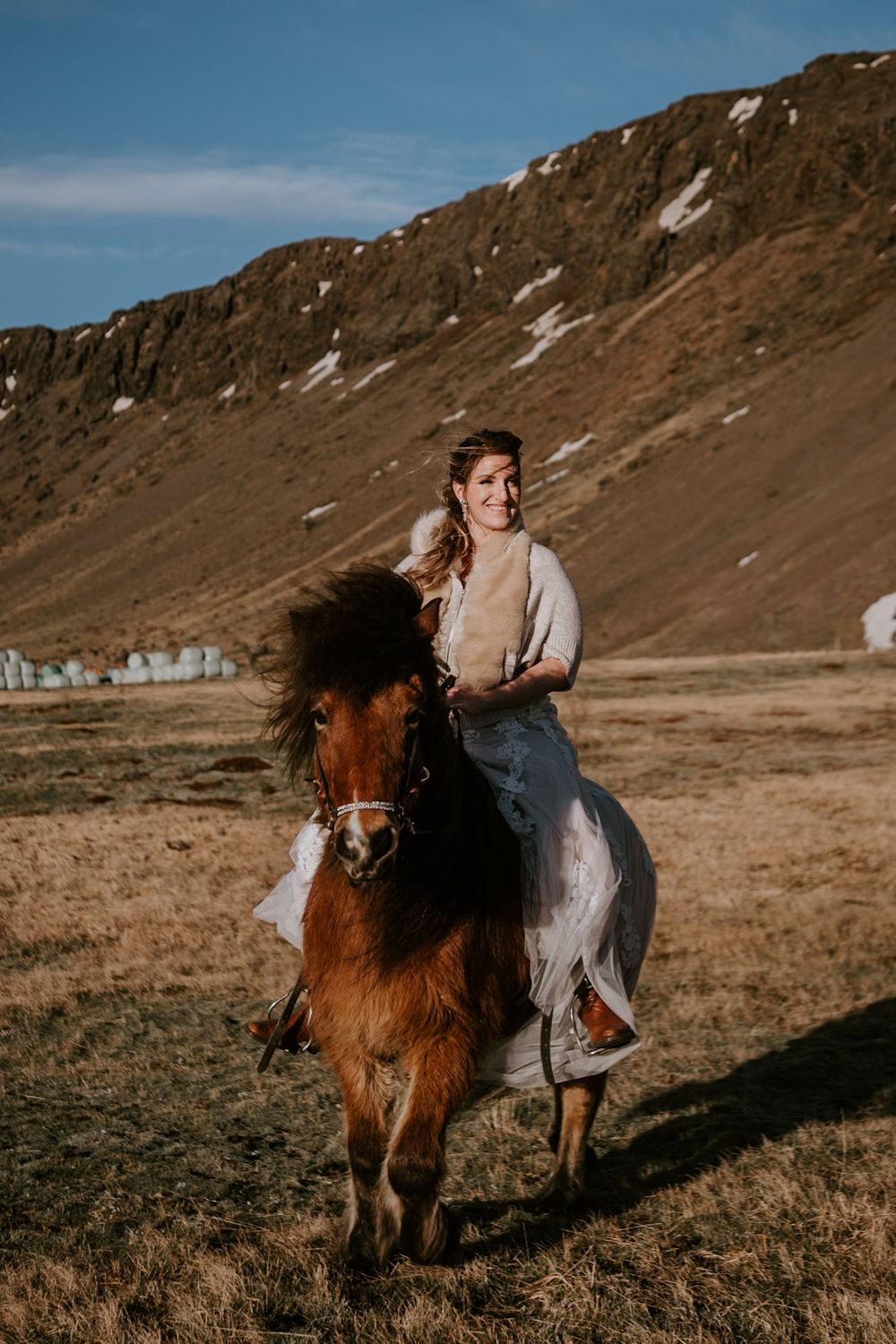 Woman in white dress riding a brown Icelandic horse in a scenic mountain landscape.