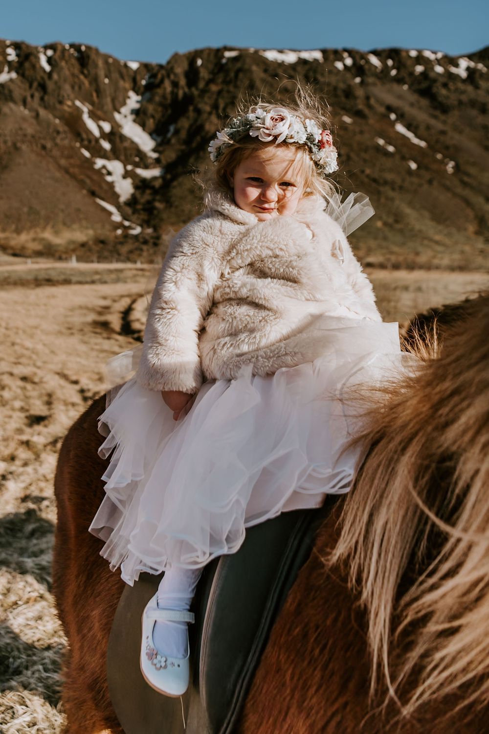 Adorable little girl in white tutu and floral crown sitting on a brown horse outdoors.
