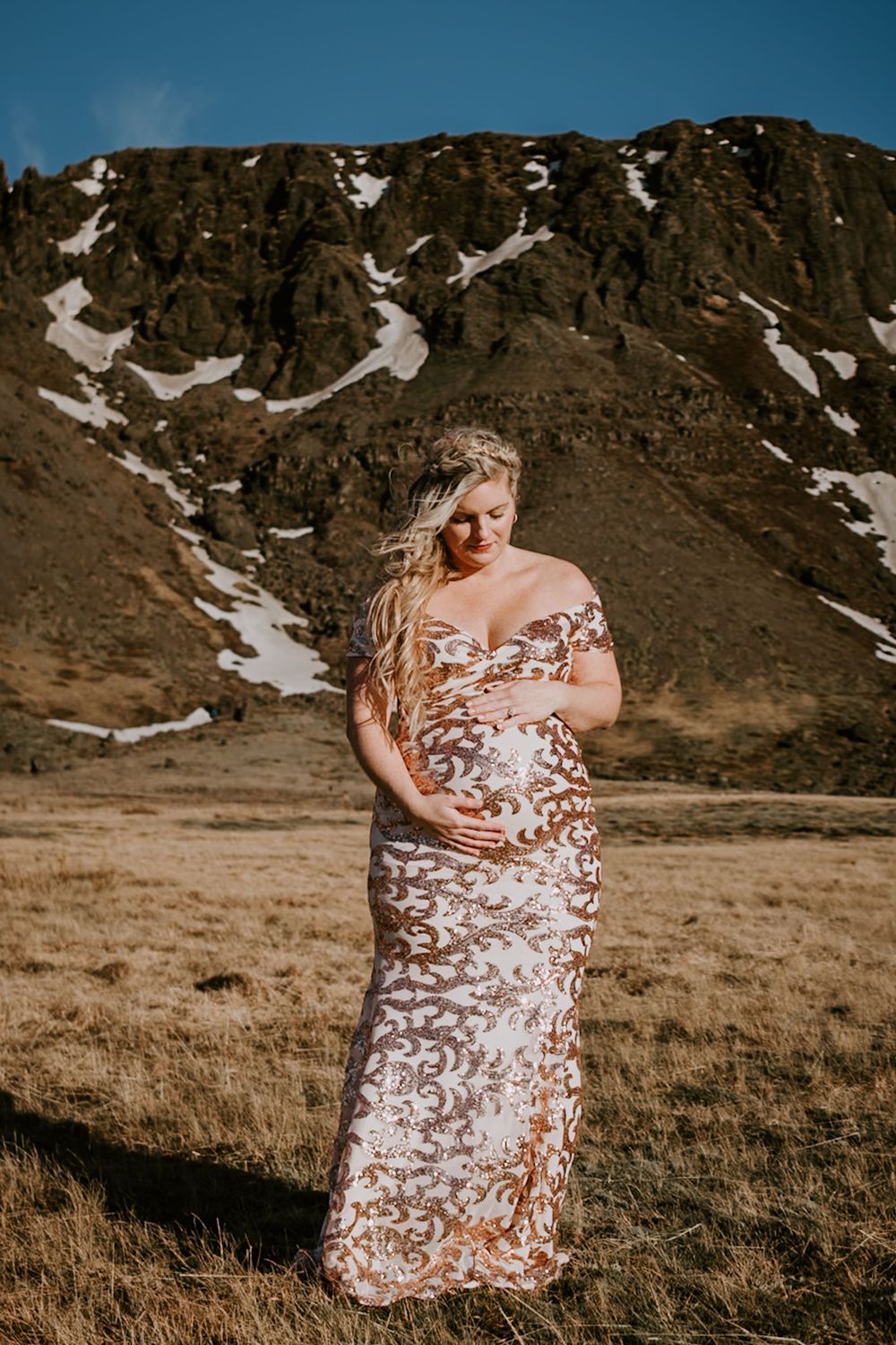 Pregnant woman in elegant lace gown posing in front of a snow-capped mountain landscape.