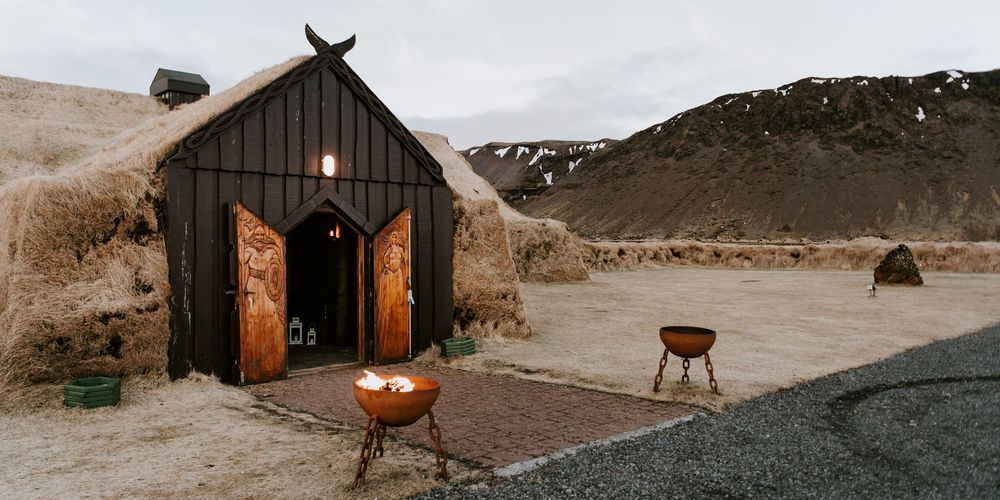 A rustic Viking-style turf house with dark wooden doors and fire pits outside in an Icelandic landscape.