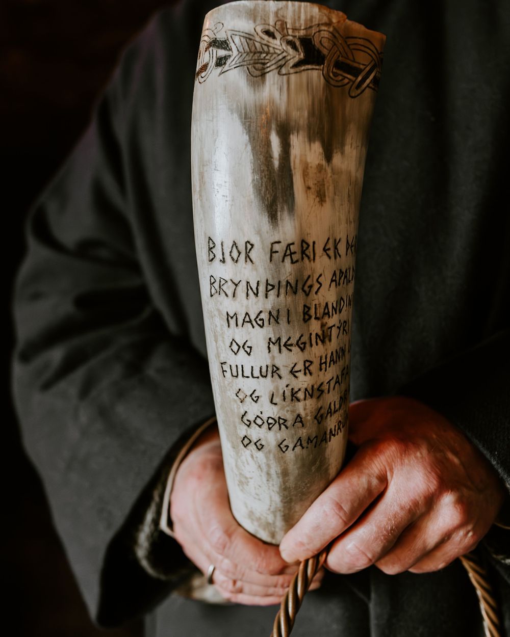 A carved drinking horn with Old Norse runic inscription held by weathered hands at a Viking ceremony.