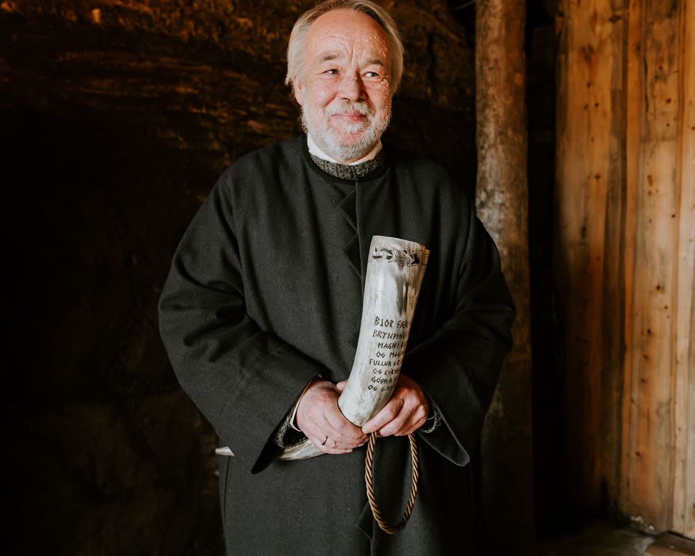 An elderly officiant in black robes holds a carved Viking drinking horn inside a rustic wooden structure.