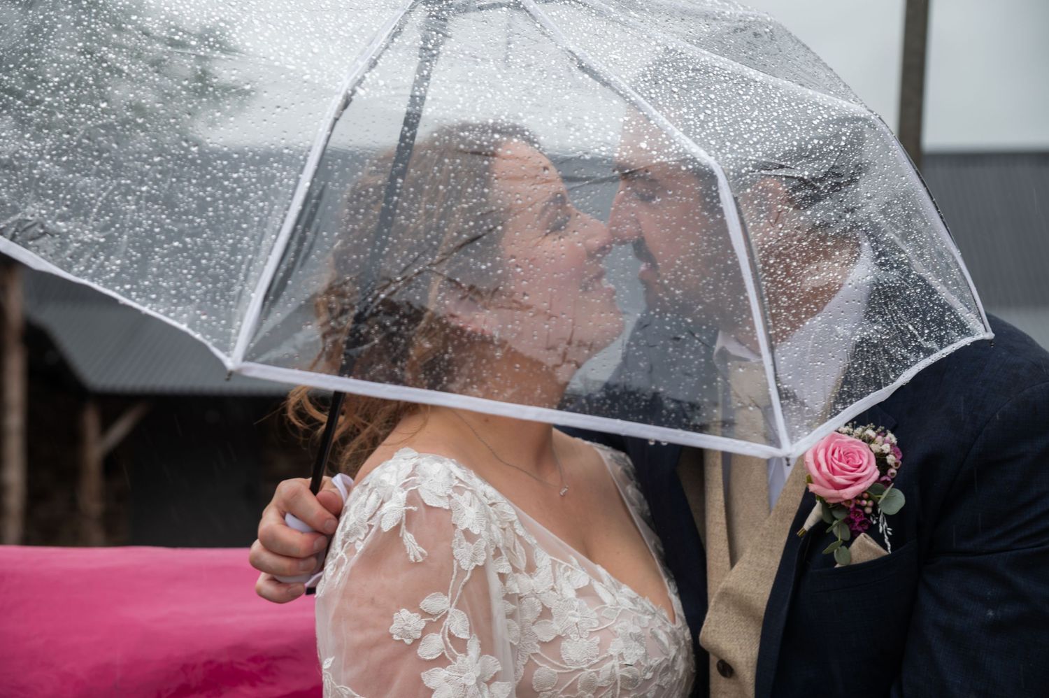 A couple stands under a clear umbrella in the rain during their wedding day celebration.