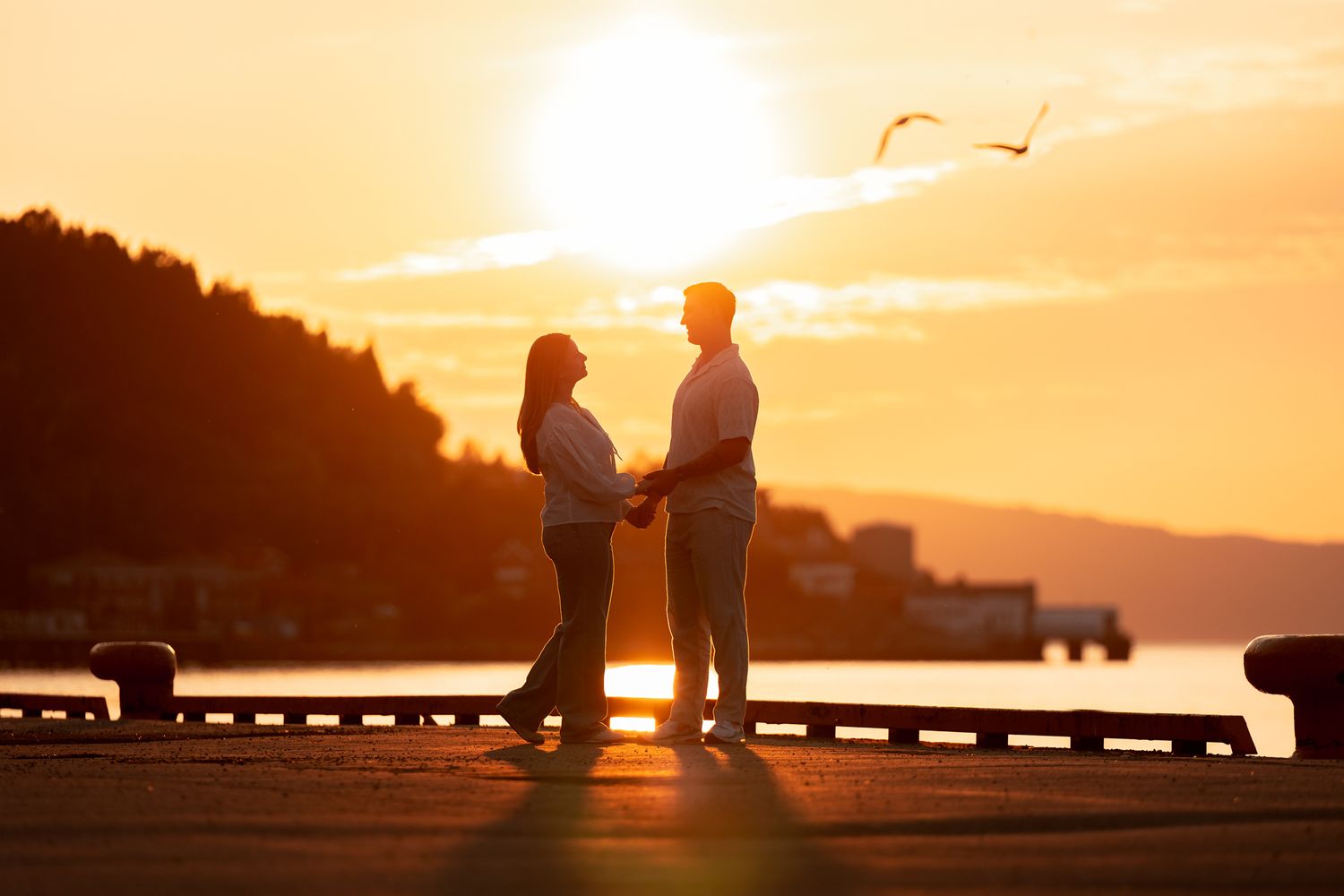 Two silhouettes stand together watching a vibrant orange sunset over a calm lake with mountains in the distance.