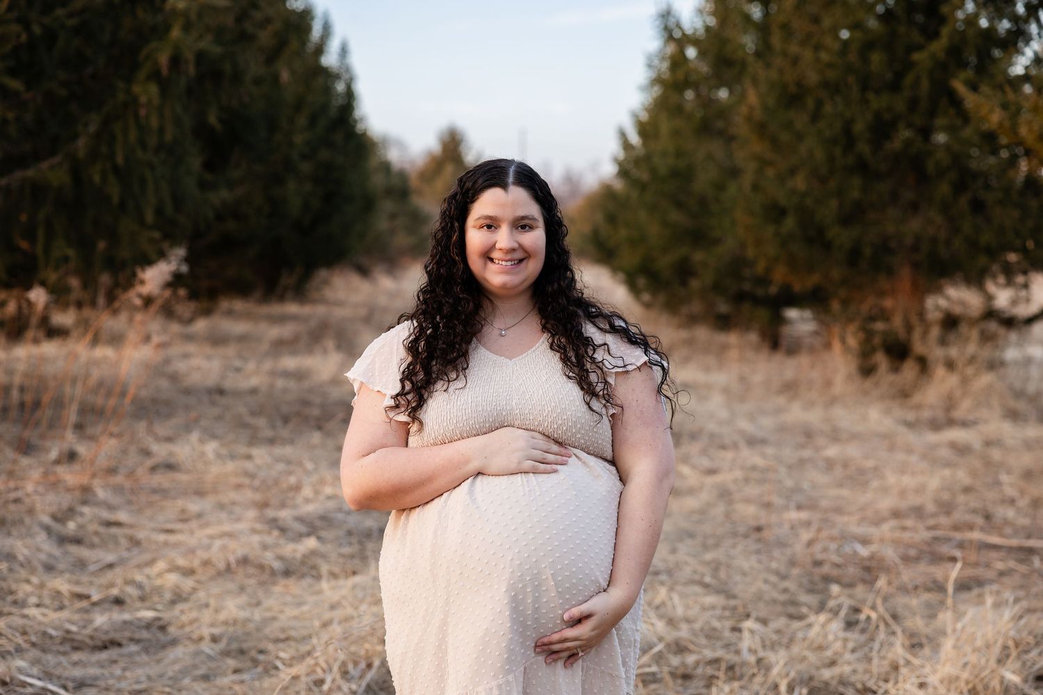 Expectant woman in a flowing cream dress stands in a field with evergreen trees in the background during golden hour.