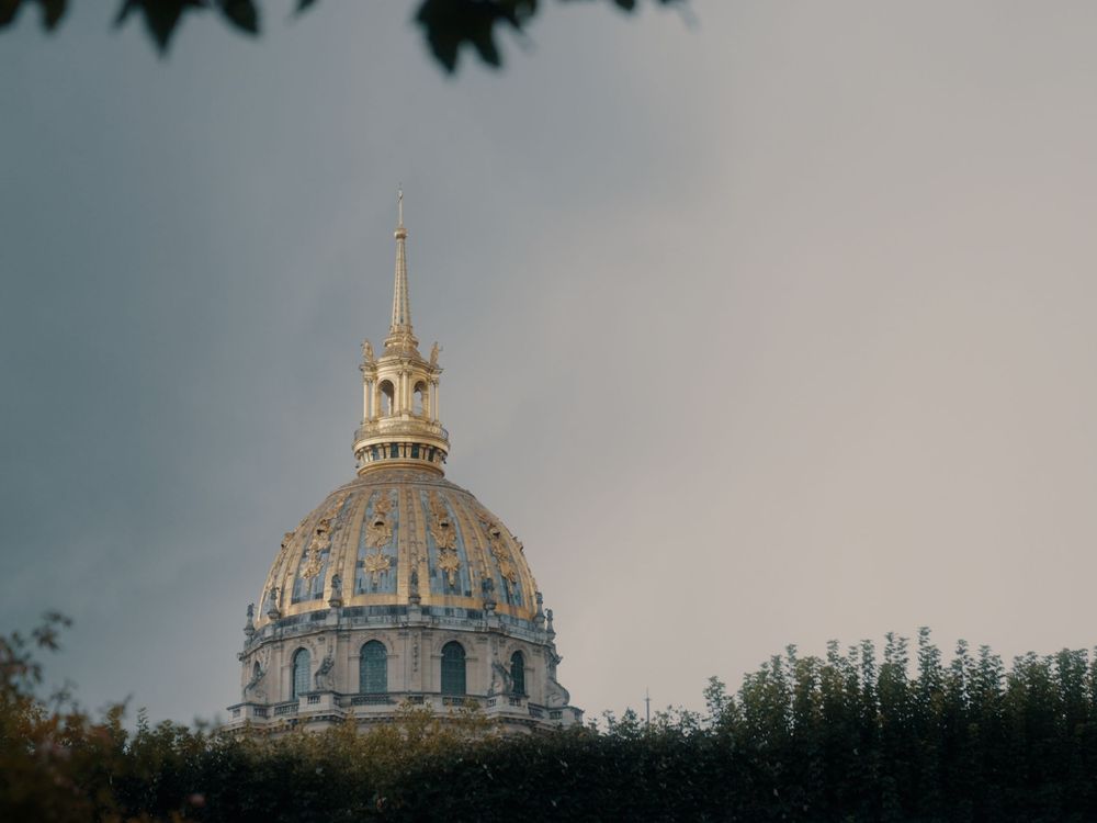 Ornate golden dome of Les Invalides in Paris rises against a cloudy sky with architectural details visible.