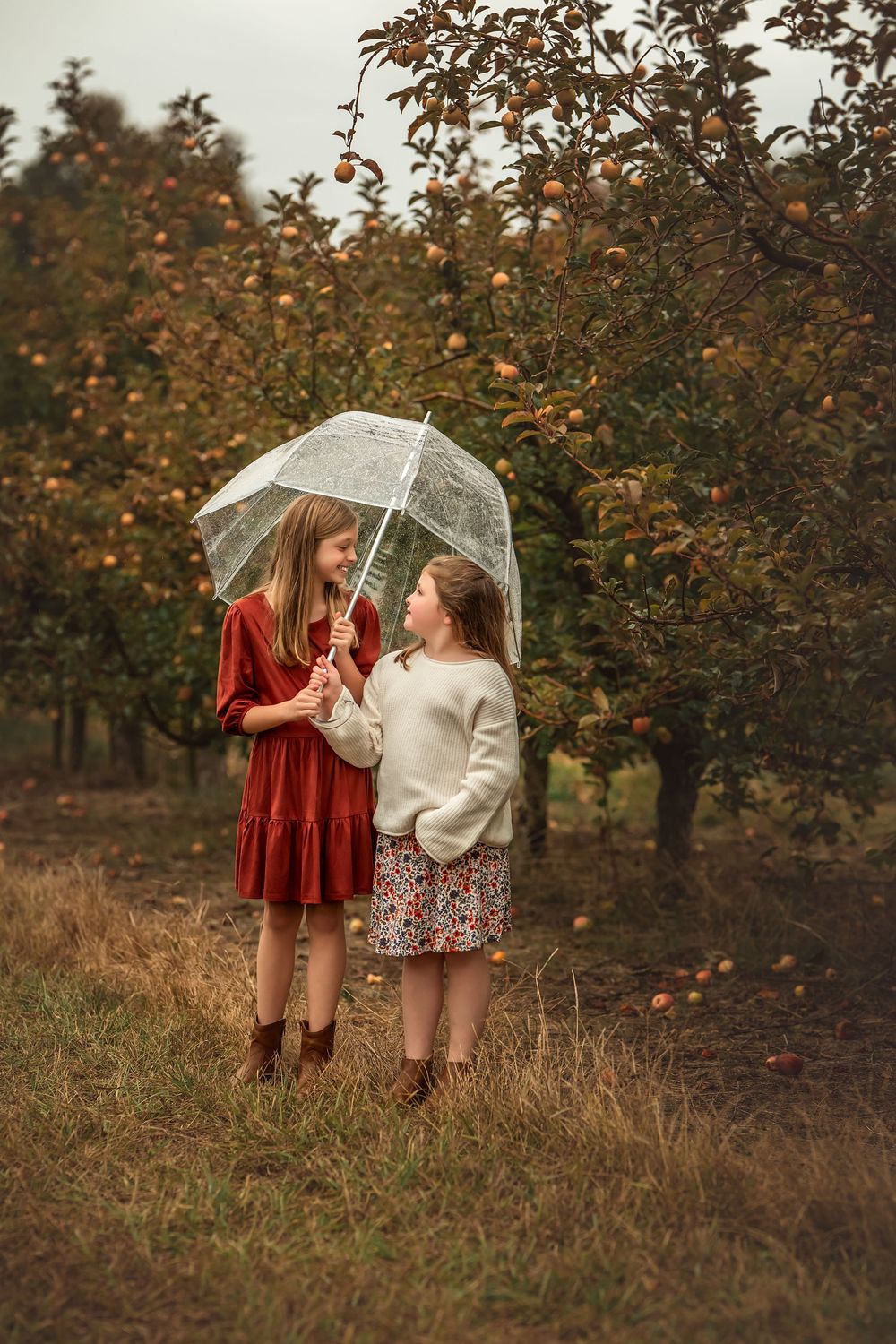 Two people in contrasting dresses share a clear umbrella among fall foliage.