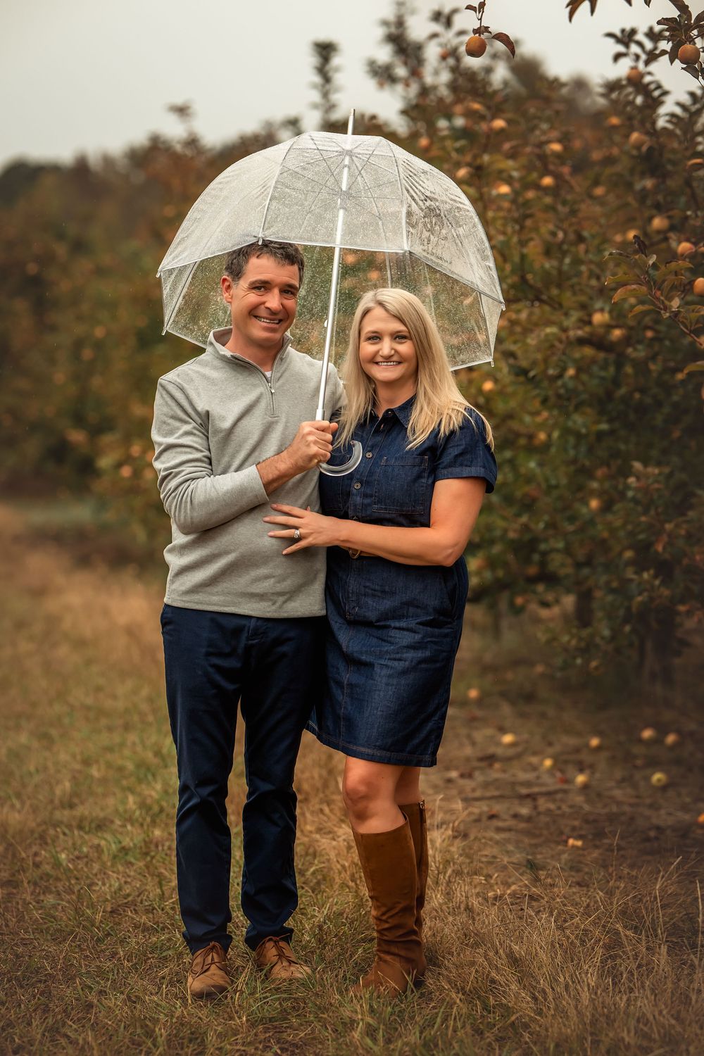 A couple shares a clear umbrella while walking through an autumn field with trees in the background.
