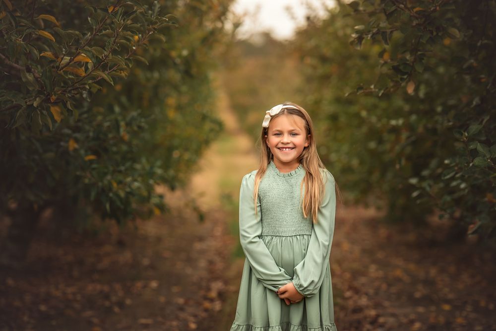 A child in a mint green dress stands between rows of fruit trees in an autumn orchard.