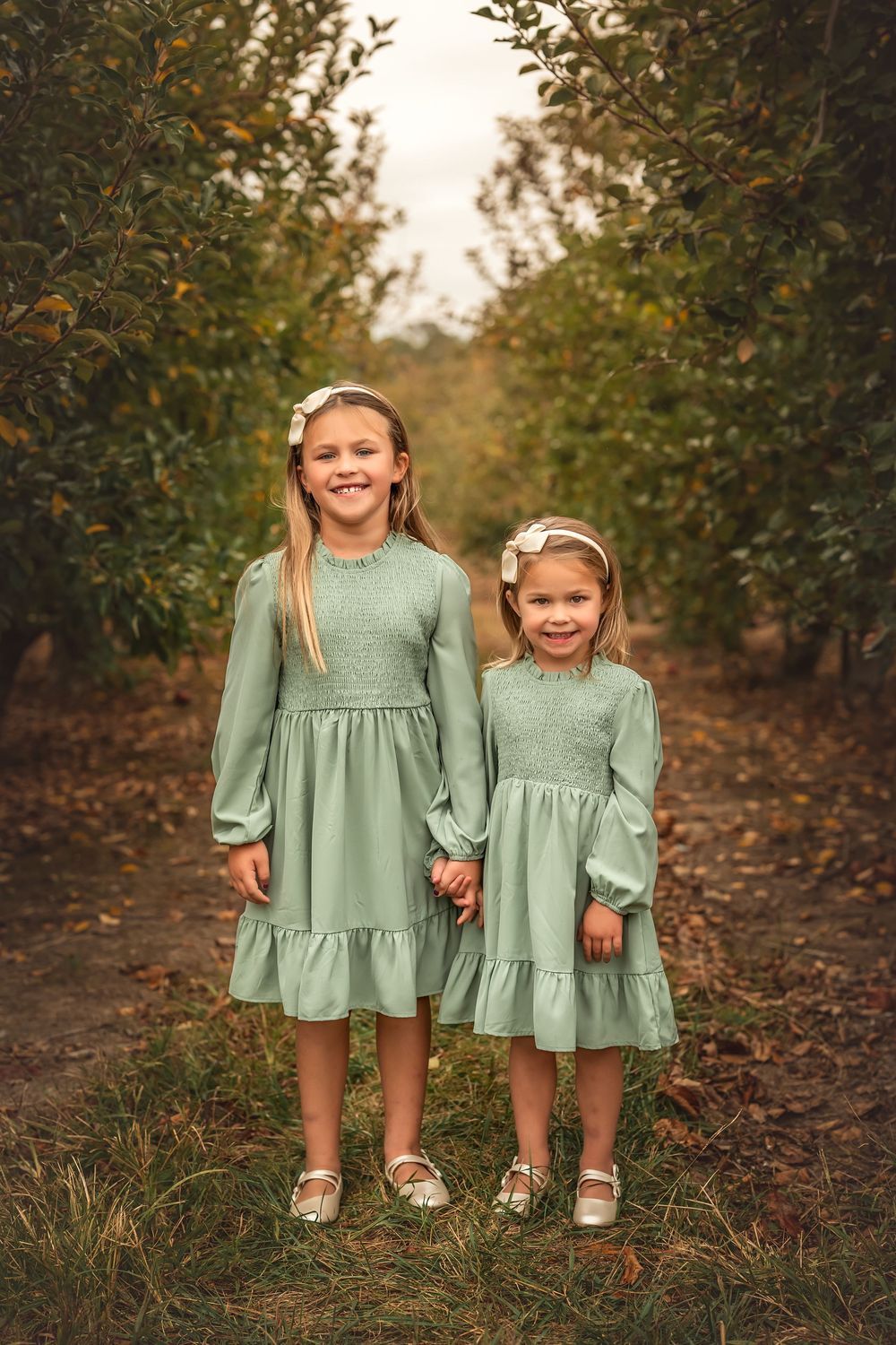 Two children in matching sage green dresses hold hands while standing in an autumn orchard.