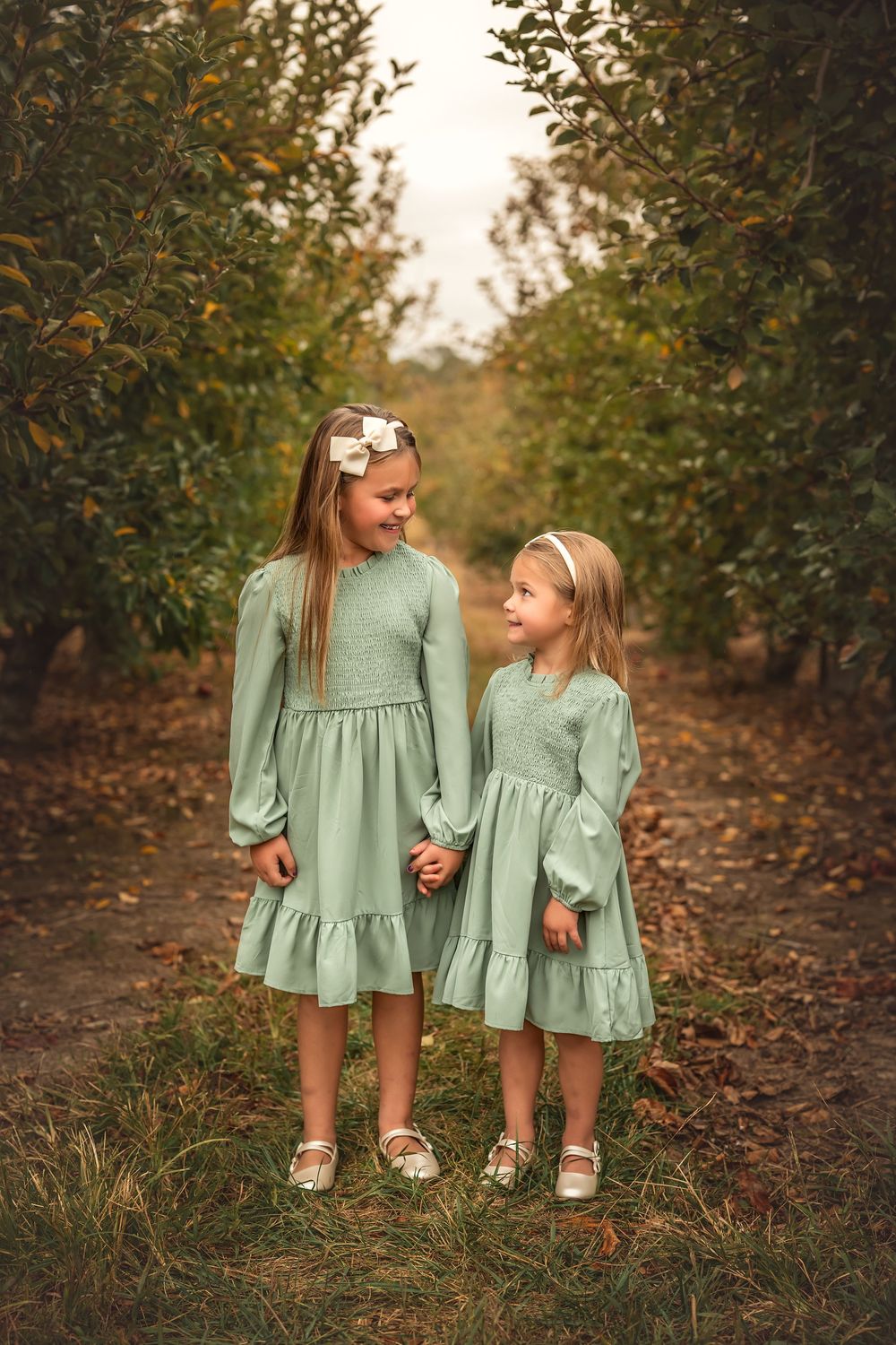 Two matching sage green dresses shown in an autumn orchard setting with fallen leaves on the ground.