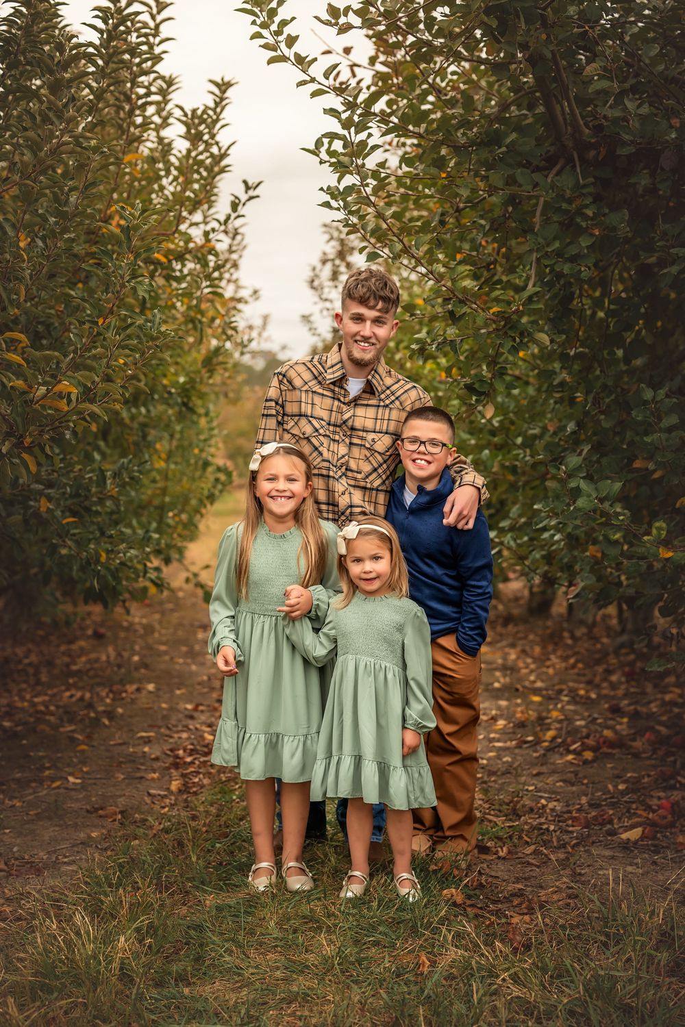A joyful autumn family photo session in an apple orchard with matching sage green dresses and brown pants.