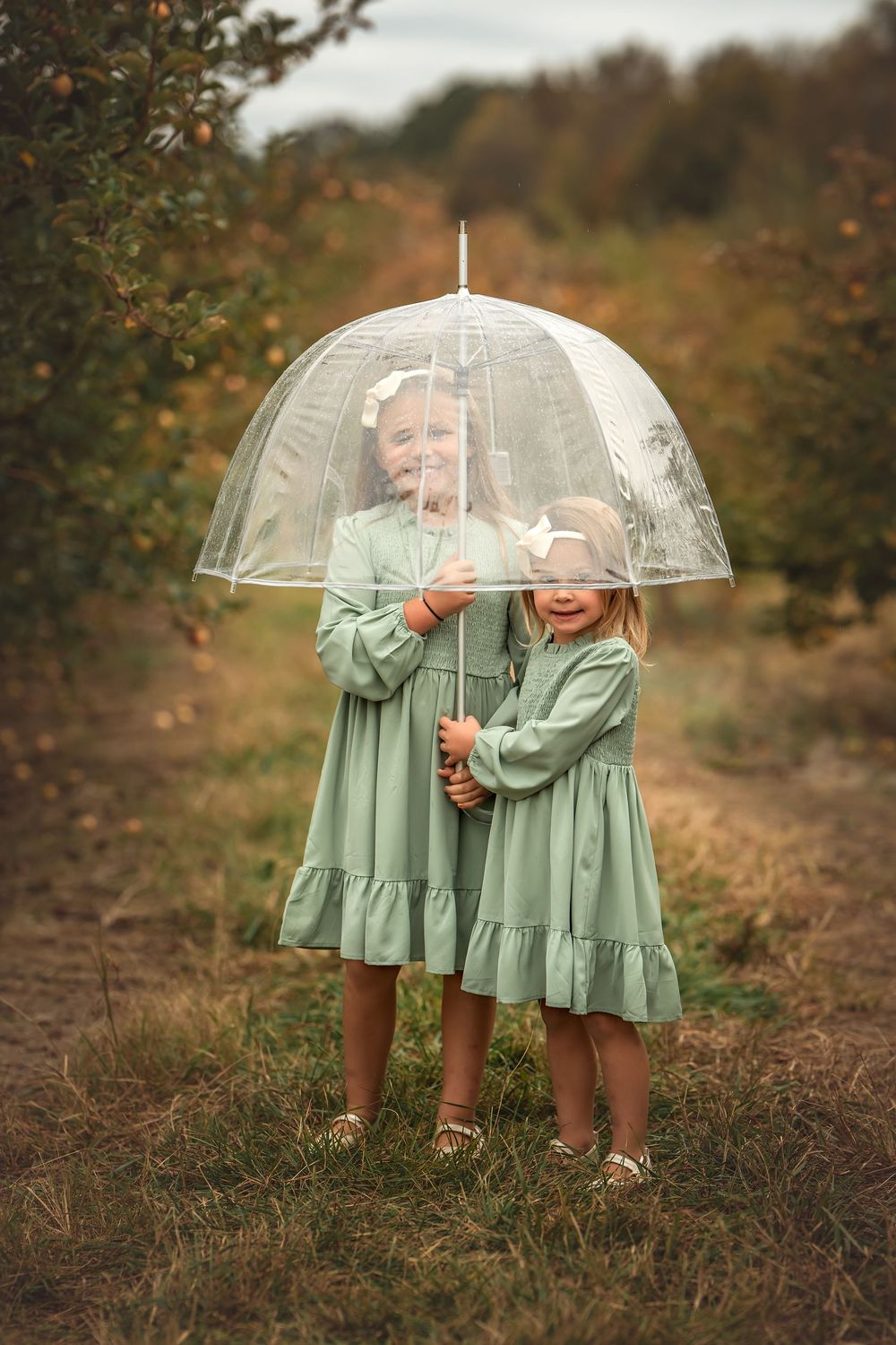 Two figures in matching mint green dresses share a clear umbrella on an autumn path.
