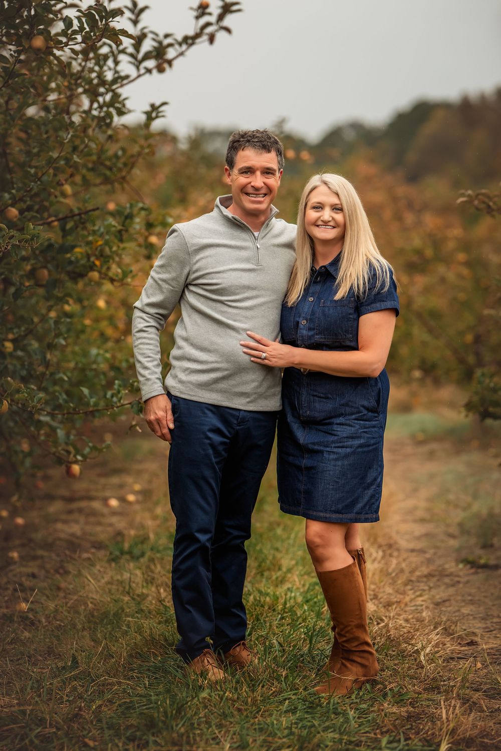 A couple poses together in an autumn apple orchard wearing coordinated navy and gray outfits.