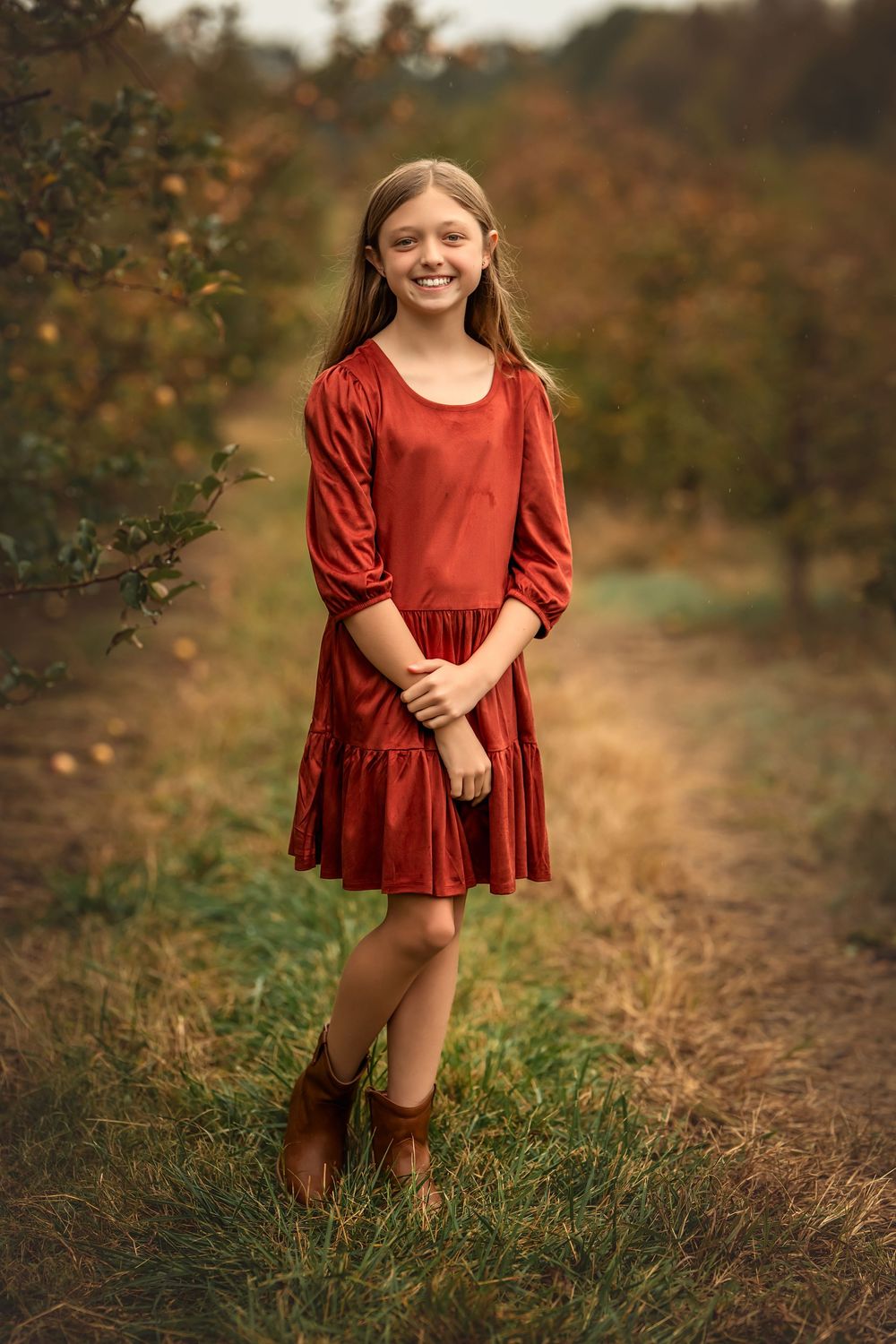 A person wearing a rust-colored velvet dress poses in an autumn field setting.