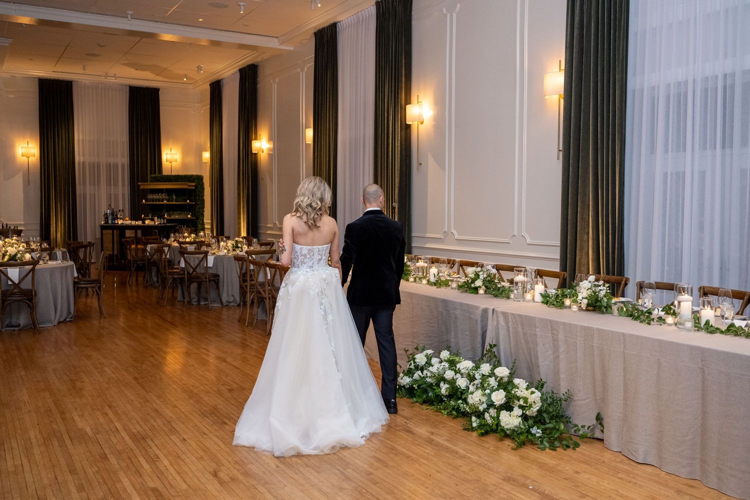 A ballroom wedding venue with hardwood floors, elegant drapes, and floral arrangements along the wall.